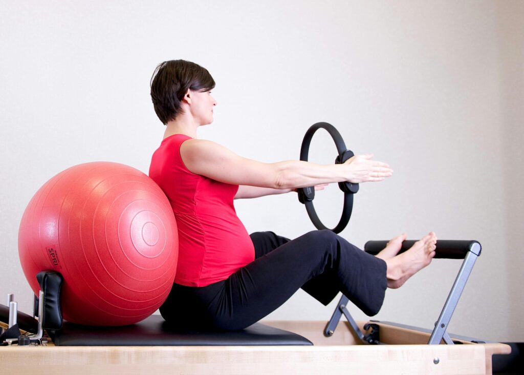 pexels-photo-1103242-1103242 A pregnant woman in a red top practicing Pilates using a fitness ring and exercise ball in a gym setting.